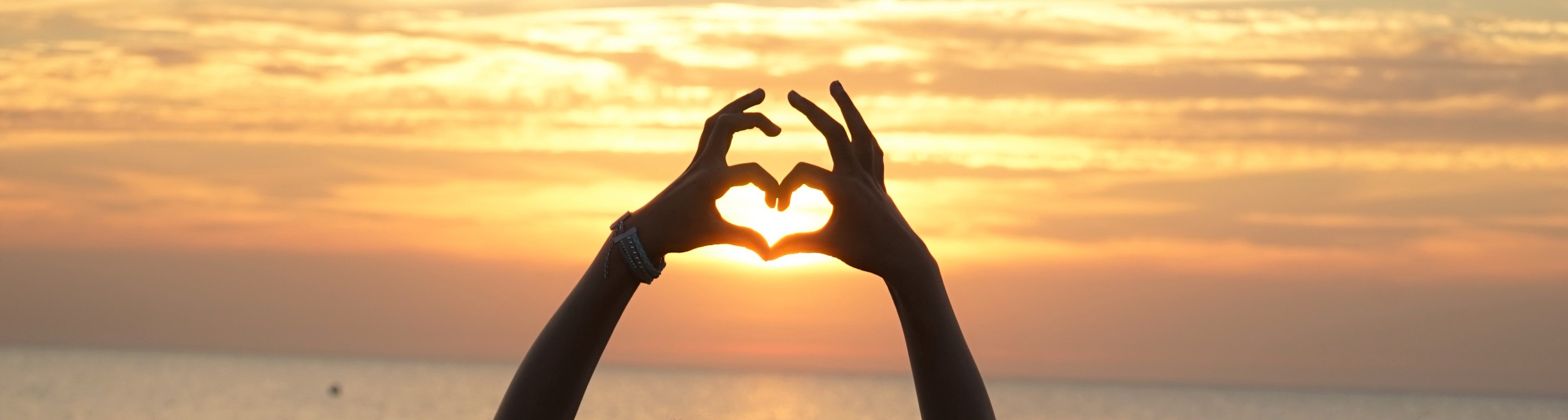 zwei Hände formen ein Herz vor einem Sonnenuntergang am Meer
